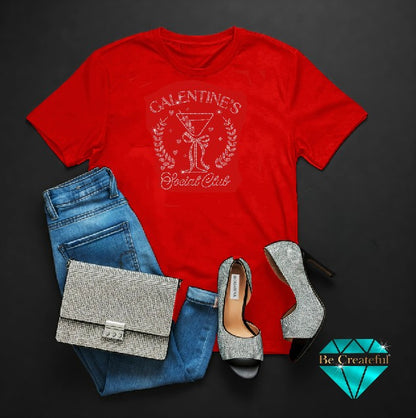 Red t-shirt with 'Galentine's Cocktail Club' design, blue jeans, gray high heels, and silver clutch on a dark background.
